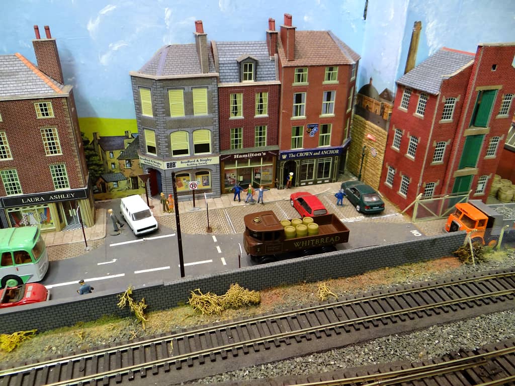Model high street with a brown steam lorry carrying barrels outside a row of shops and a pub, people on the pavement, and railway tracks in the foreground.
