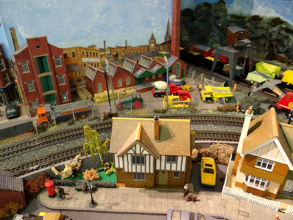 Looking down on a model Tudor-style house and garden beside a curving railway, with a builder’s yard full of lorries and machinery and a quarry beyond.