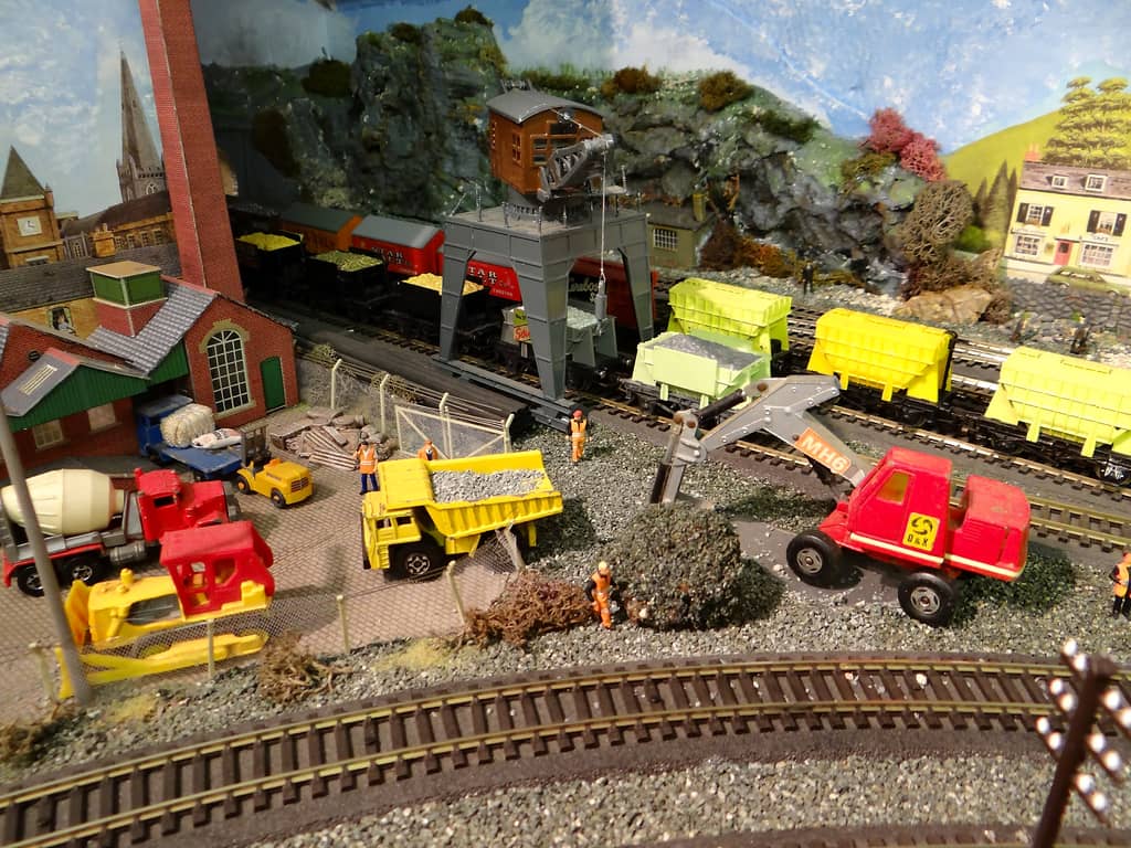 A model builder’s yard with a yellow dump truck, bulldozer and cement mixer, a red excavator by a heap of stone, and a grey gantry crane over lines of wagons in the quarry sidings.