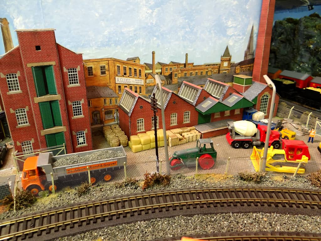 A model builder’s yard with an orange tipper, a green road roller, a cement mixer and a yellow bulldozer behind a wire fence, in front of a red-brick mill and a sawtooth-roofed factory.