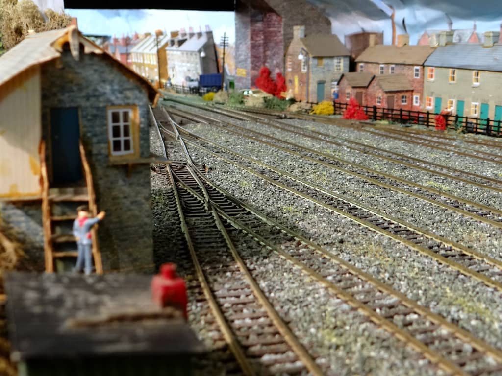 Low view across empty model sidings and points to brick terraced houses with blue-green doors, with a grey stone hut, wooden steps and a small figure on the left.