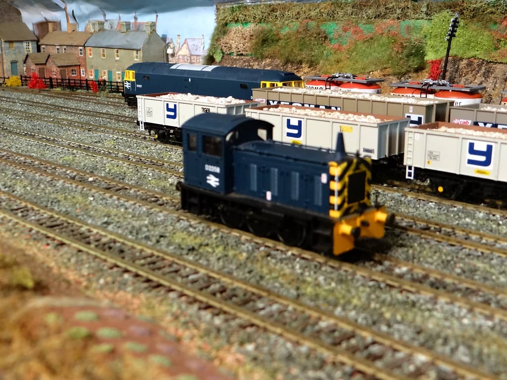 A small dark blue shunting locomotive with black and yellow striped ends in front of white wagons full of stone, with a larger blue diesel and houses behind.