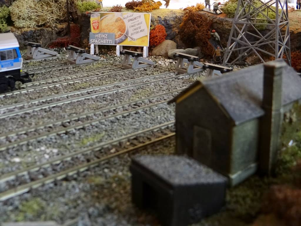 A model poster hoarding advertising Heinz Cooked Spaghetti behind a row of buffer stops, with tiny workmen in a trench and a lattice pylon to the right.