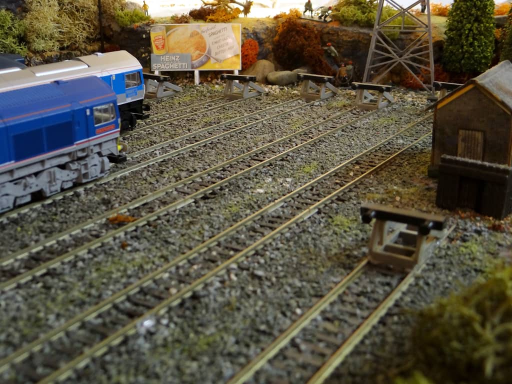 Model sidings running to a row of buffer stops under a Heinz spaghetti poster, with two blue diesels on the left and a lattice pylon and a small hut on the right.