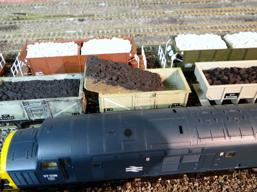Top-down view of a blue diesel locomotive’s roof and cab beside open wagons of coal and of white stone, one coal load tipped up, with the number 37 038 on the cab side.