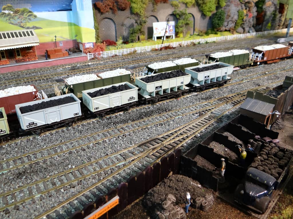 Rows of grey coal wagons and wagons of white stone on yard tracks, with wooden coal bins, sacks, workmen and a dark blue lorry in front and a station platform behind.