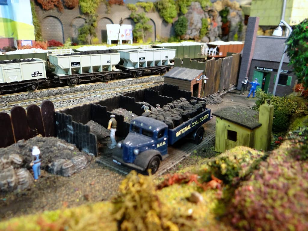 A model coal yard with a dark blue NCB lorry loaded with sacks, men in wooden coal bins, a small brick office with a green door signed Ivor Lump, and grey coal wagons behind a fence.