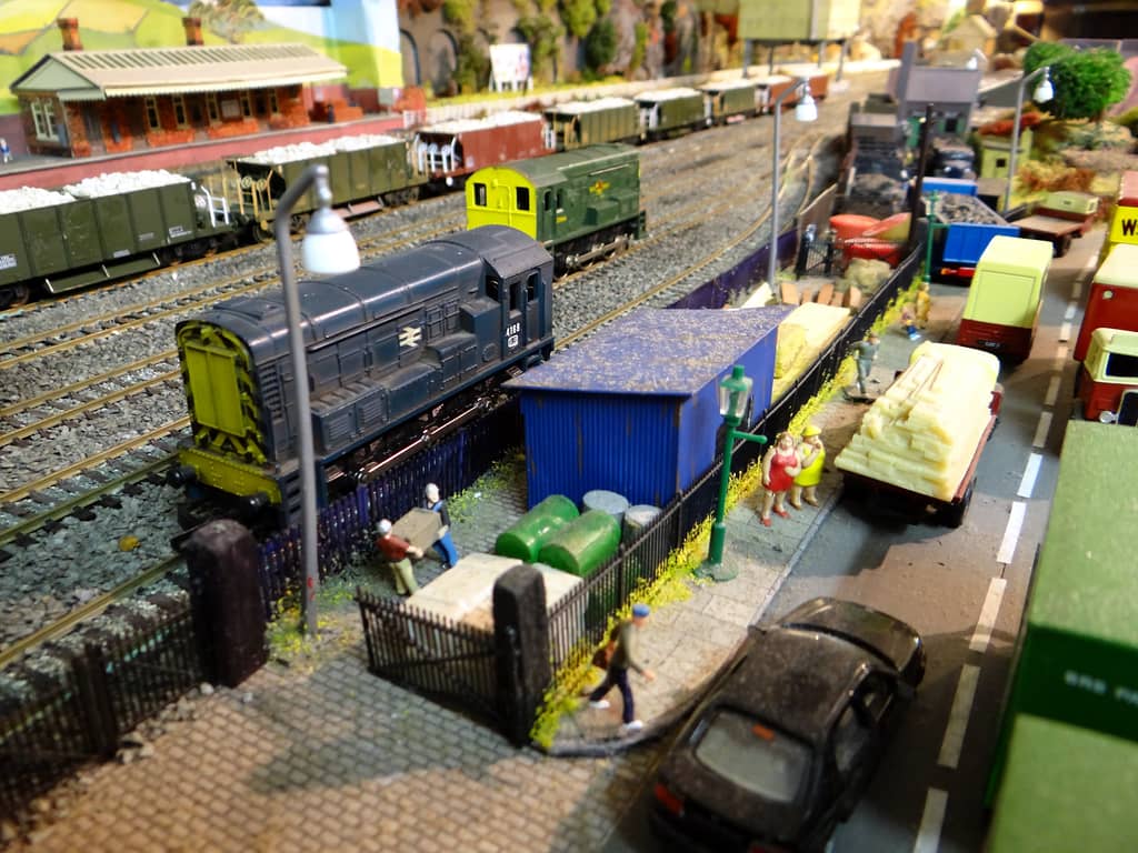A dark blue shunting engine and a green one on yard tracks, with a blue hut, green oil drums, figures on a pavement, parked lorries and a dark car on the road.