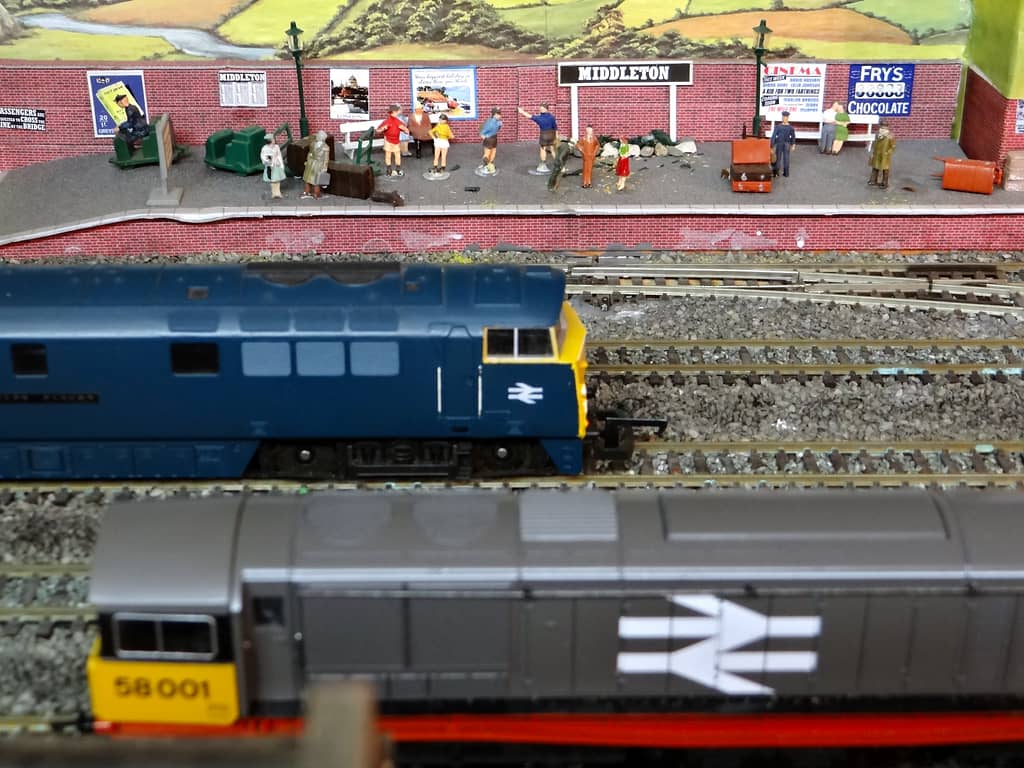 A model station platform with a “MIDDLETON” nameboard, posters and many small figures against a brick wall, with a blue diesel and a grey diesel numbered 58001 in front.