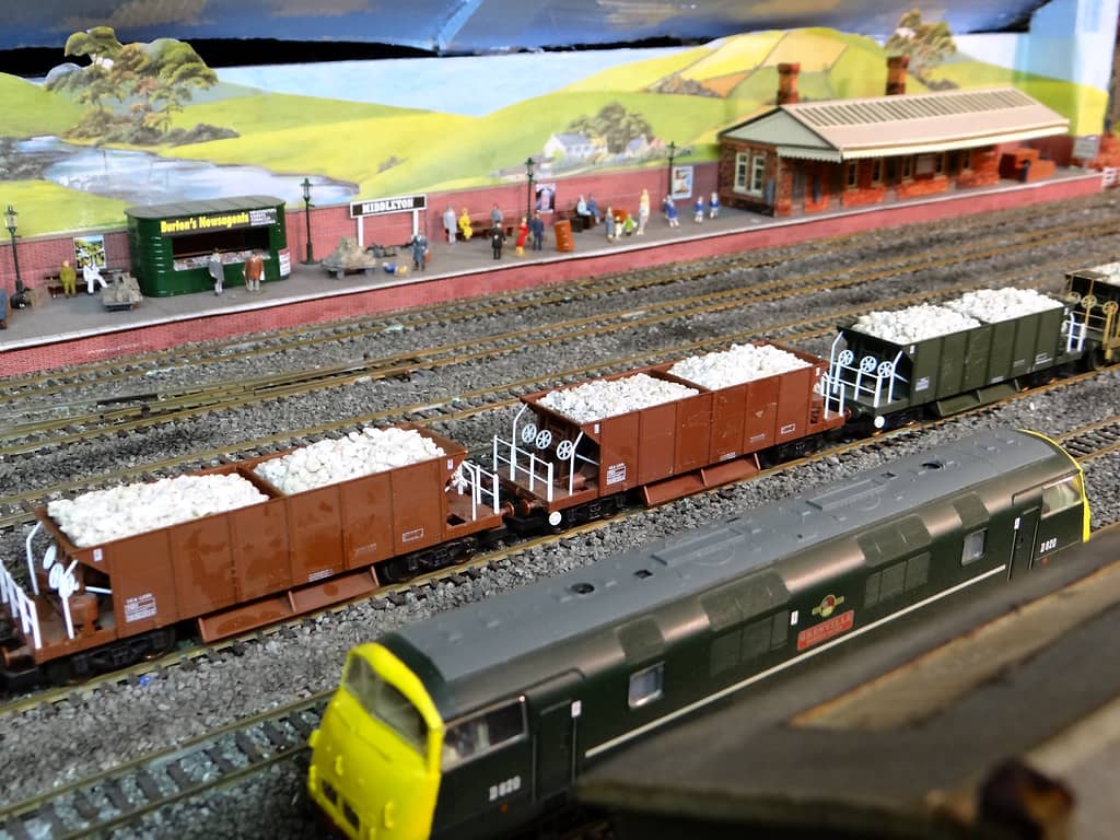 A green diesel with a yellow end in the foreground, brown and green wagons loaded with white stone, and behind them a platform with a green kiosk, nameboard and station building.