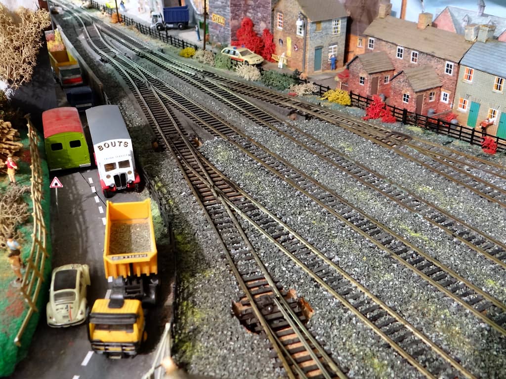 A road on the left with a yellow tipper lorry of gravel, a cream Beetle, a white van and a green van beside a fan of railway points, with brick terraced houses on the right.