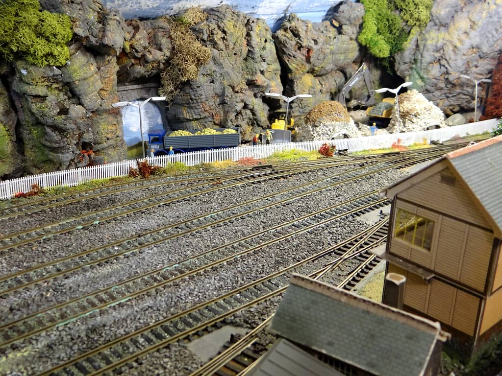 A model quarry of grey rock faces with a stone-arched tunnel, a blue lorry of yellow stone, workmen in hard hats, a grey excavator, heaps of white stone and a signal box in front.
