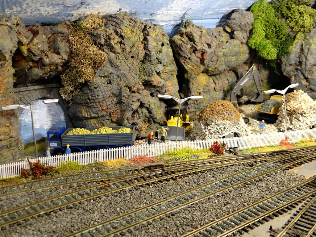 Grey rock faces with a tunnel mouth, a blue and grey lorry full of yellow stone, workmen in hard hats behind a white picket fence, a bulldozer and a grey excavator.
